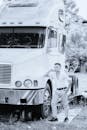 A monochrome image of a man leaning on a large white truck outdoors.
