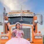 A young woman in a pink gown smiles in front of a vibrant orange truck under a clear blue sky.